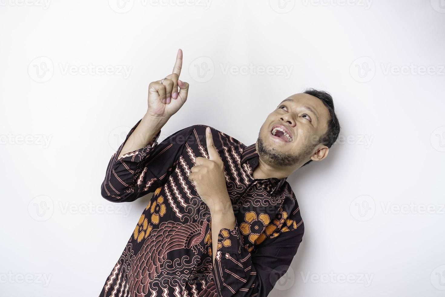 Excited Asian man wearing batik shirt pointing at the copy space on top of him, isolated by white background photo