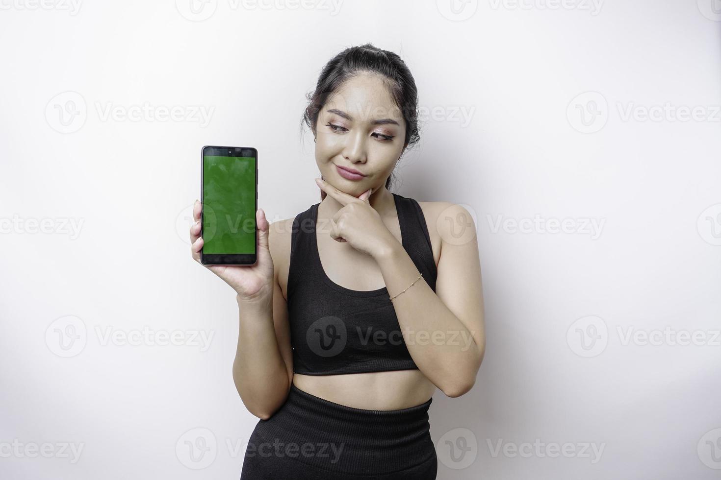 una joven deportista pensativa lleva ropa deportiva mientras se sostiene la barbilla y muestra una pantalla verde en su teléfono, aislada de fondo blanco foto