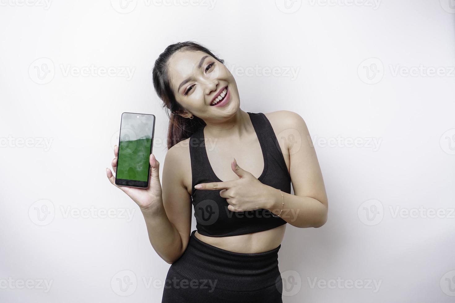 A portrait of a sporty Asian woman wearing a sportswear and showing green screen on her phone, isolated by white background photo