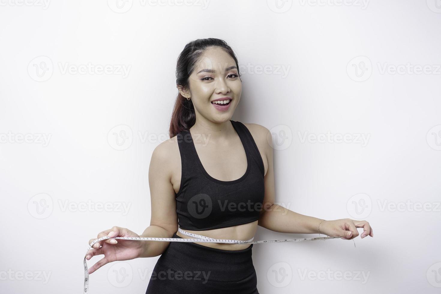 Portrait of cheerful young Asian woman in sportswear measuring waist with tape, feeling happy about losing weight at home. Healthy lifestyle, sports and slimming diet concept photo