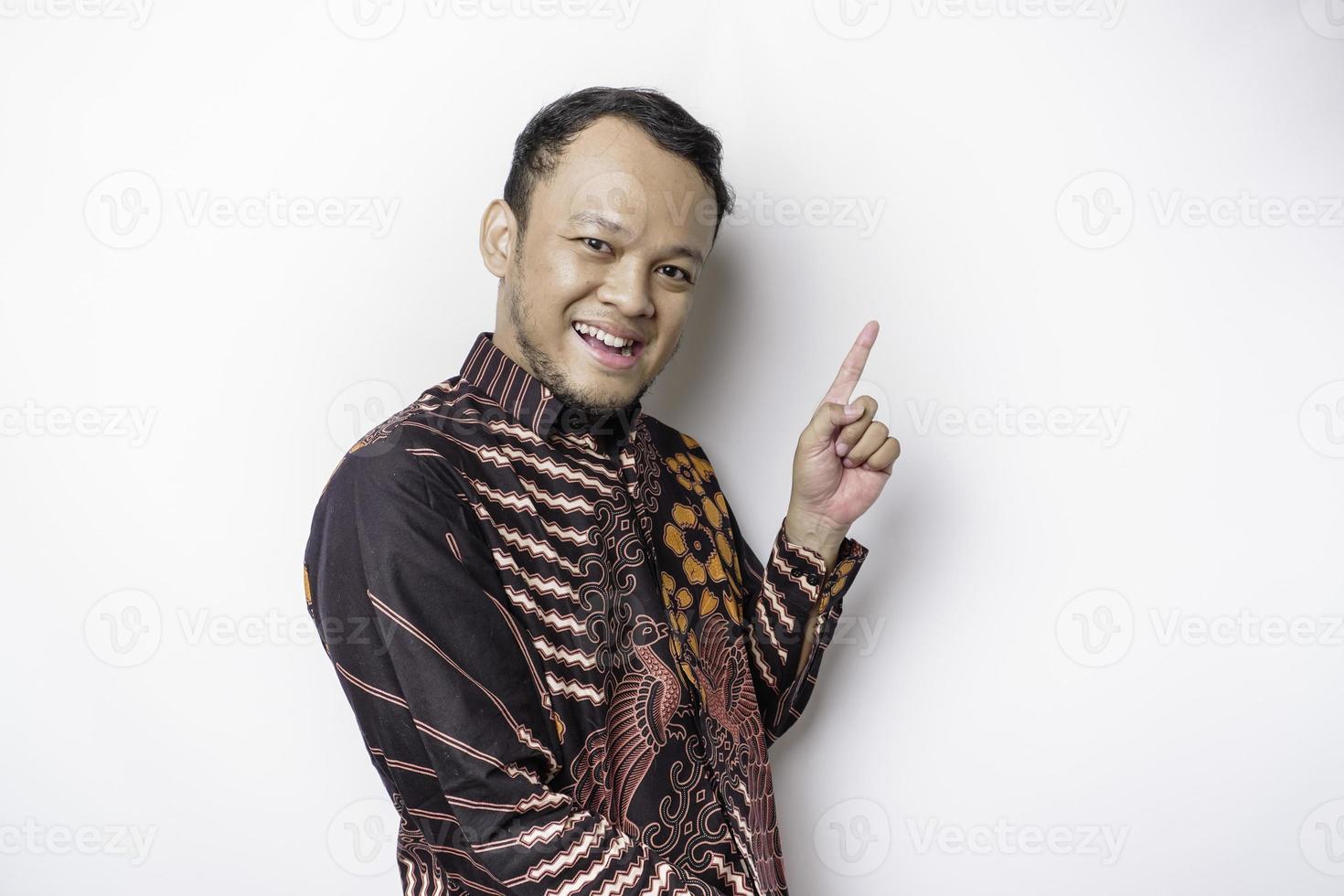 Excited Asian man wearing batik shirt pointing at the copy space on top of him, isolated by white background photo