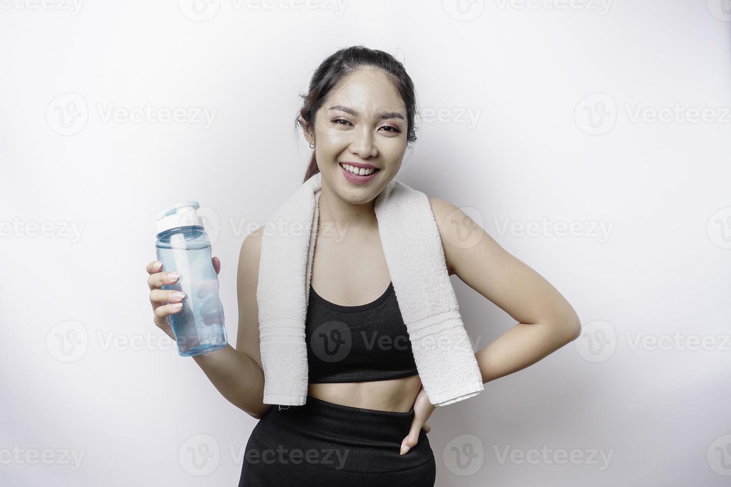 mujer asiática deportiva posando con una toalla en el hombro y sosteniendo una botella de agua, sonriendo y relajándose después del entrenamiento foto