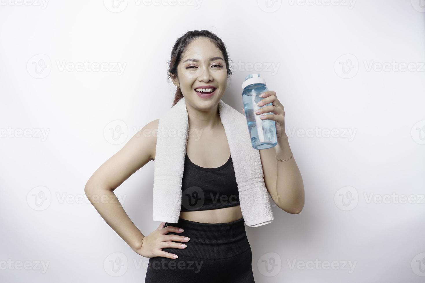 mujer asiática deportiva posando con una toalla en el hombro y sosteniendo una botella de agua, sonriendo y relajándose después del entrenamiento foto