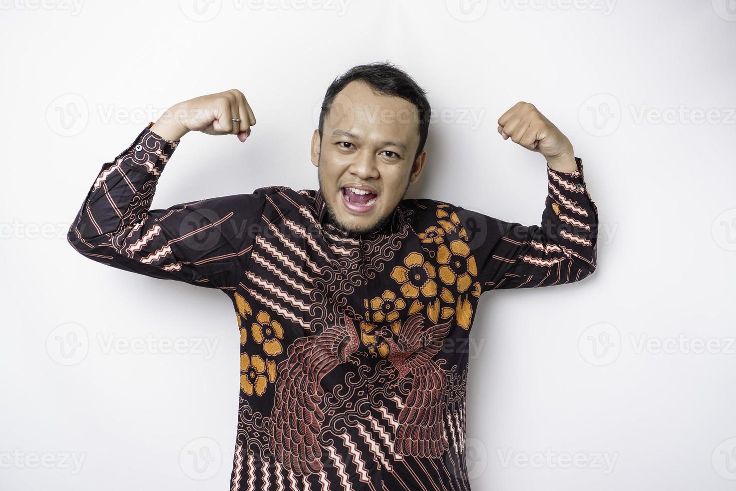 Excited Asian man wearing batik shirt showing strong gesture by lifting his arms and muscles smiling proudly photo