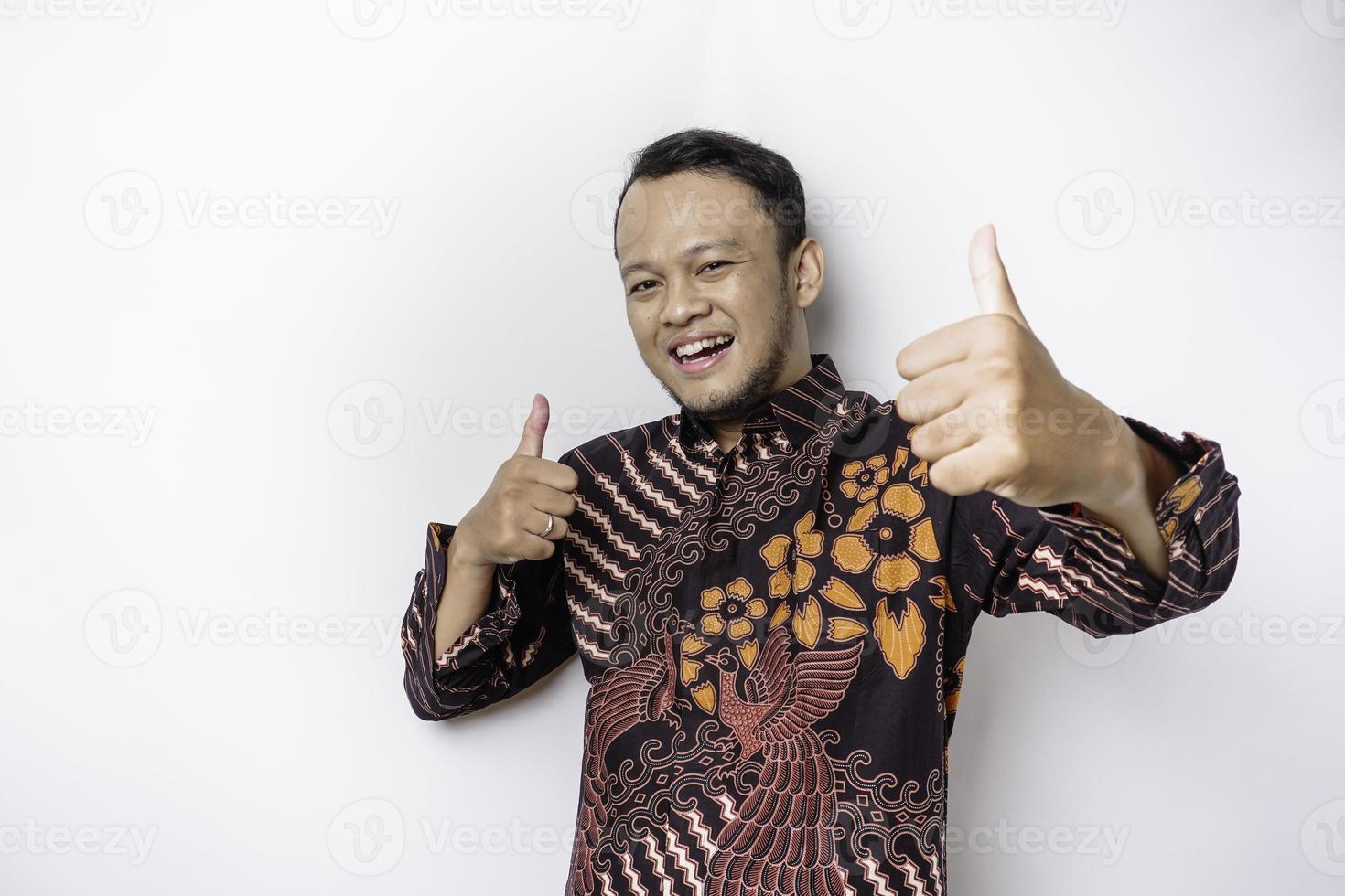 Excited Asian man wears batik shirt, gives thumbs up hand gesture of approval, isolated by white background photo