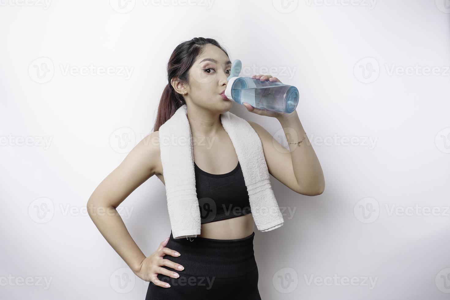Sportive Asian woman posing with a towel on her shoulder and drinking from a bottle of water, smiling and relaxing after workout photo