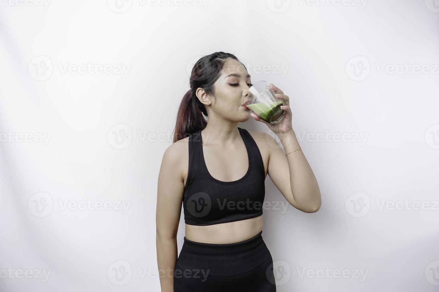 Joyful sporty Asian woman wearing sportswear is drinking a glass of tasty green smoothie, isolated on white background. photo