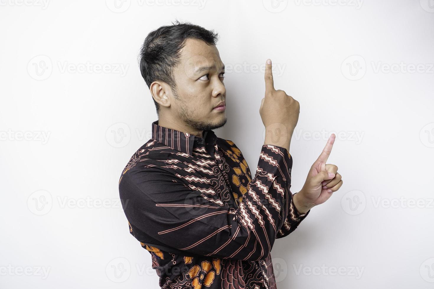 Serious Asian man wearing batik shirt pointing at the copy space on top of him, isolated by white background photo