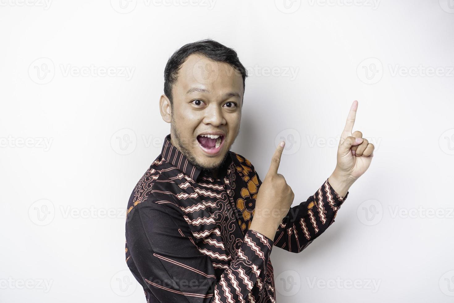 Excited Asian man wearing batik shirt pointing at the copy space on top of him, isolated by white background photo