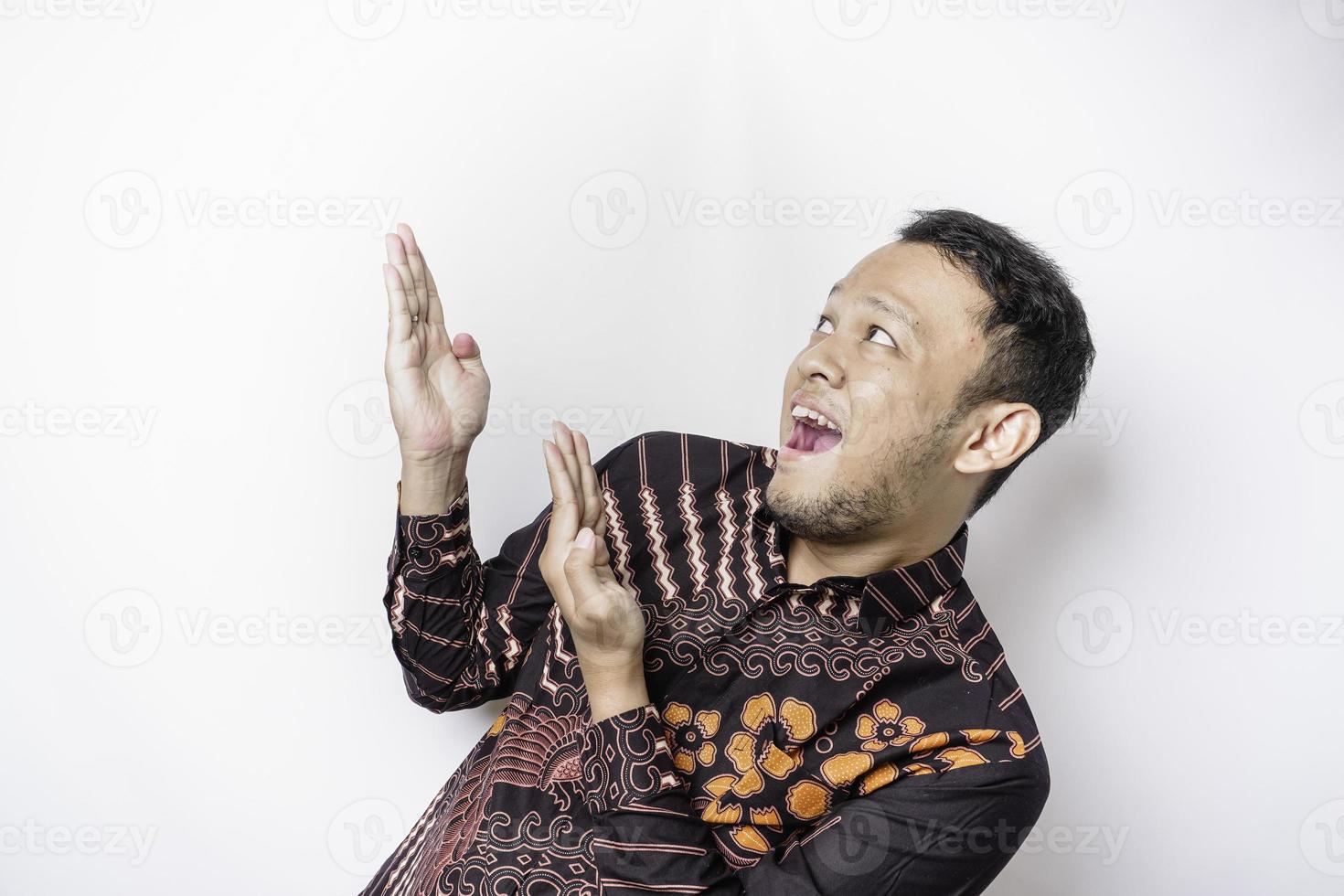 Excited Asian man wearing batik shirt pointing at the copy space on top of him, isolated by white background photo