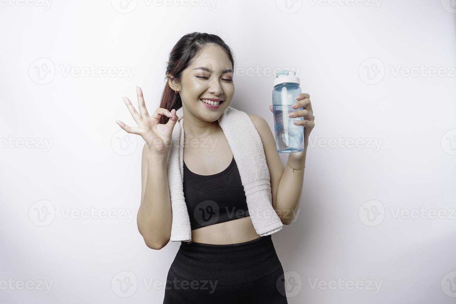 mujer asiática deportiva sonriente posando con una toalla en el hombro y sosteniendo una botella de agua mientras gesticulaba con el dedo, relajándose después del entrenamiento foto