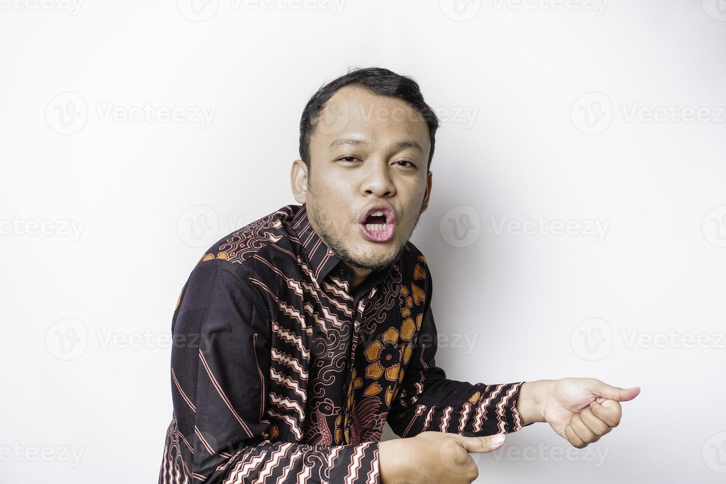 Shocked Asian man wearing batik shirt pointing at the copy space beside him, isolated by white background photo