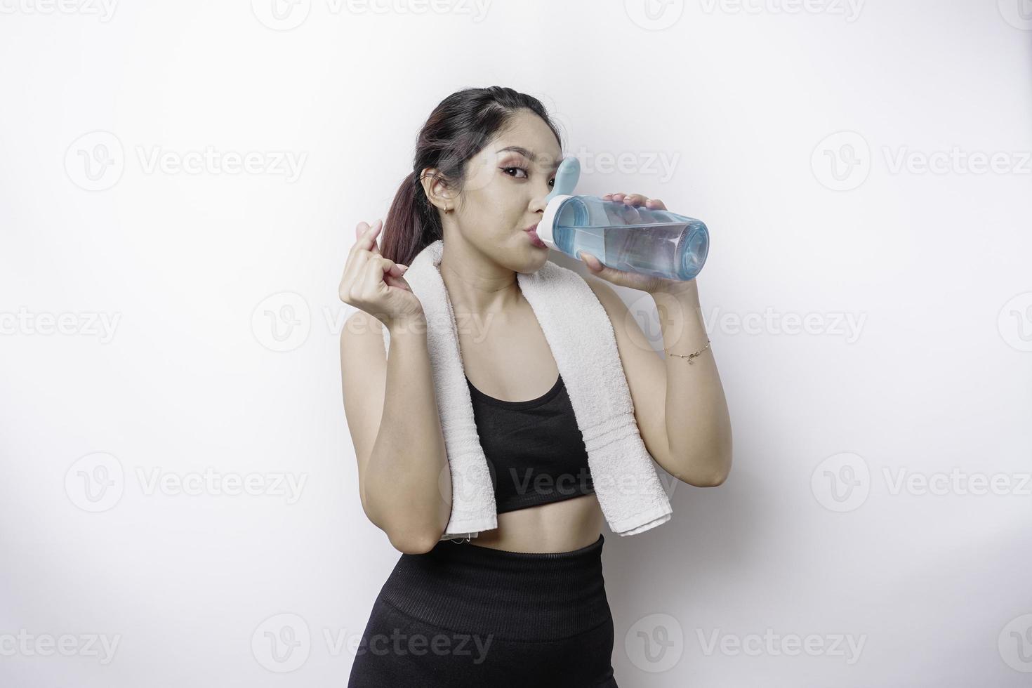 Sportive Asian woman posing with a towel on her shoulder and drinking from a bottle of water, smiling and relaxing after workout photo