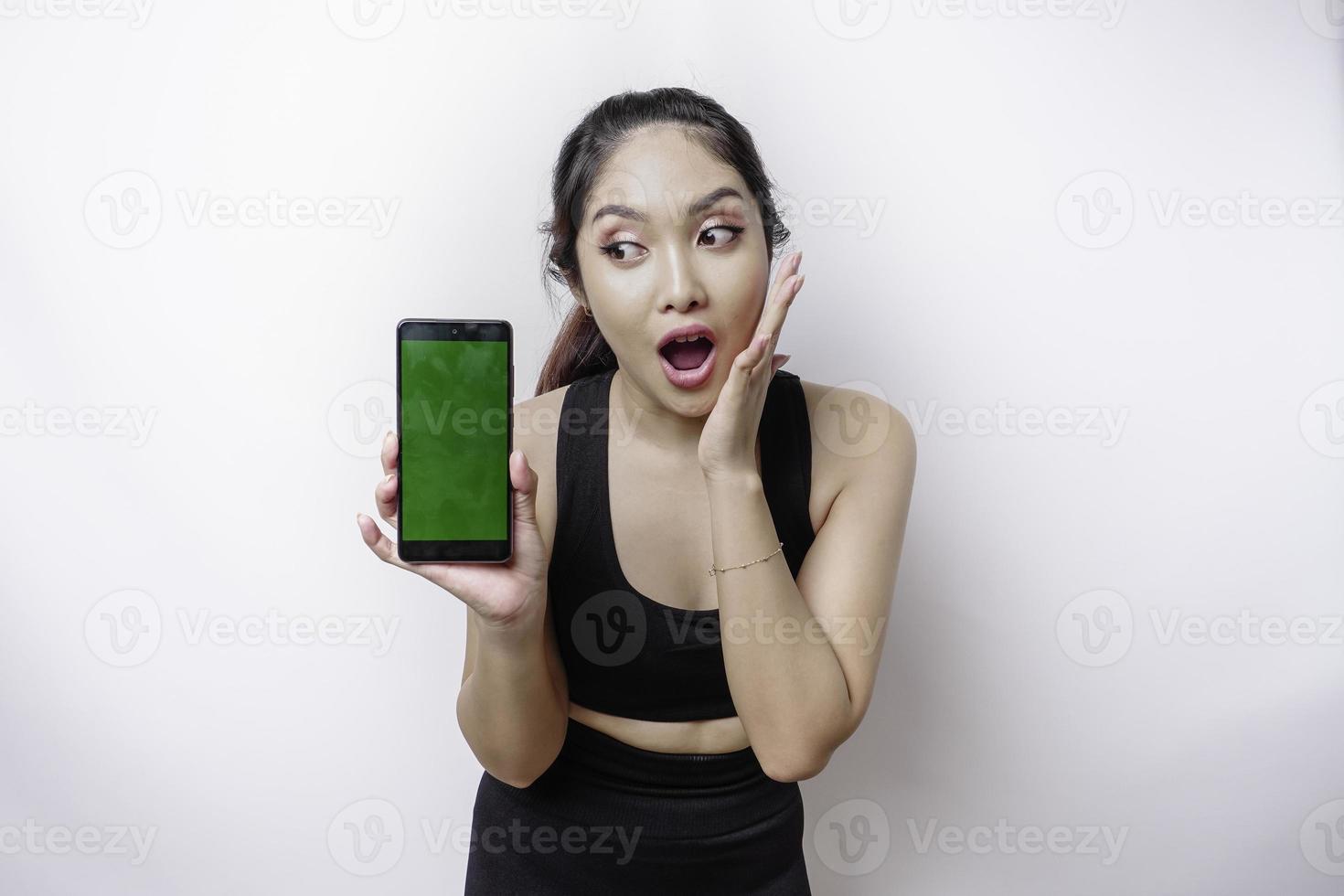 Shocked sporty Asian woman wearing sportswear and showing green screen on her phone, isolated by white background photo