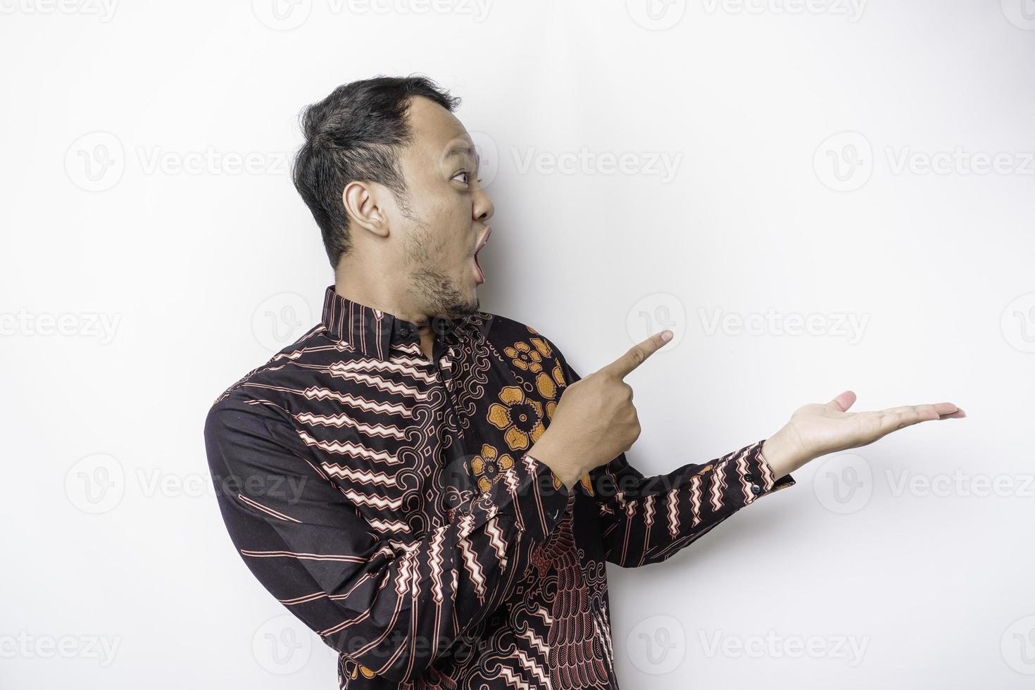 Shocked Asian man wearing batik shirt pointing at the copy space beside him, isolated by white background photo