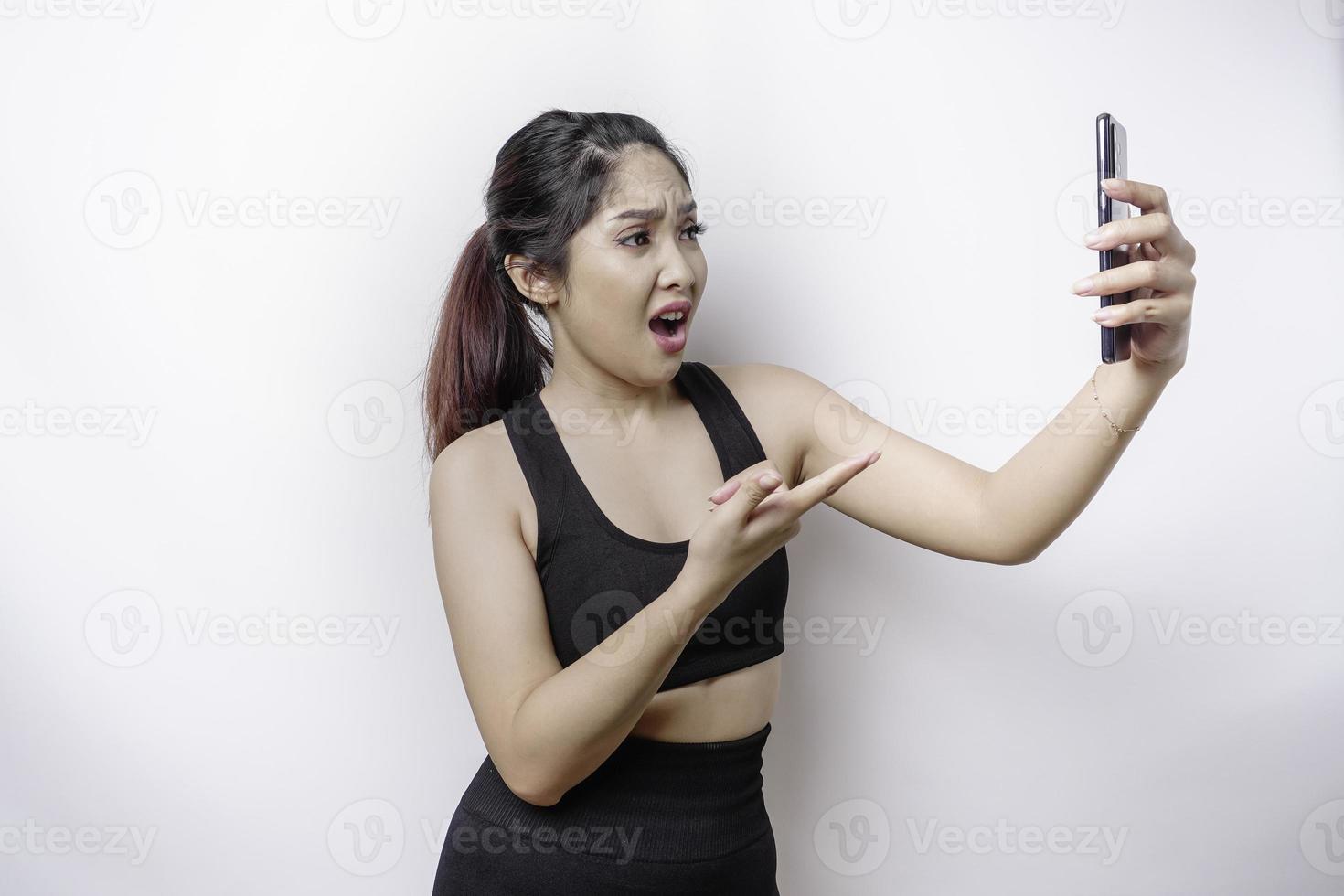 A dissatisfied sporty Asian woman looks disgruntled wearing sportswear irritated face expressions holding her phone photo