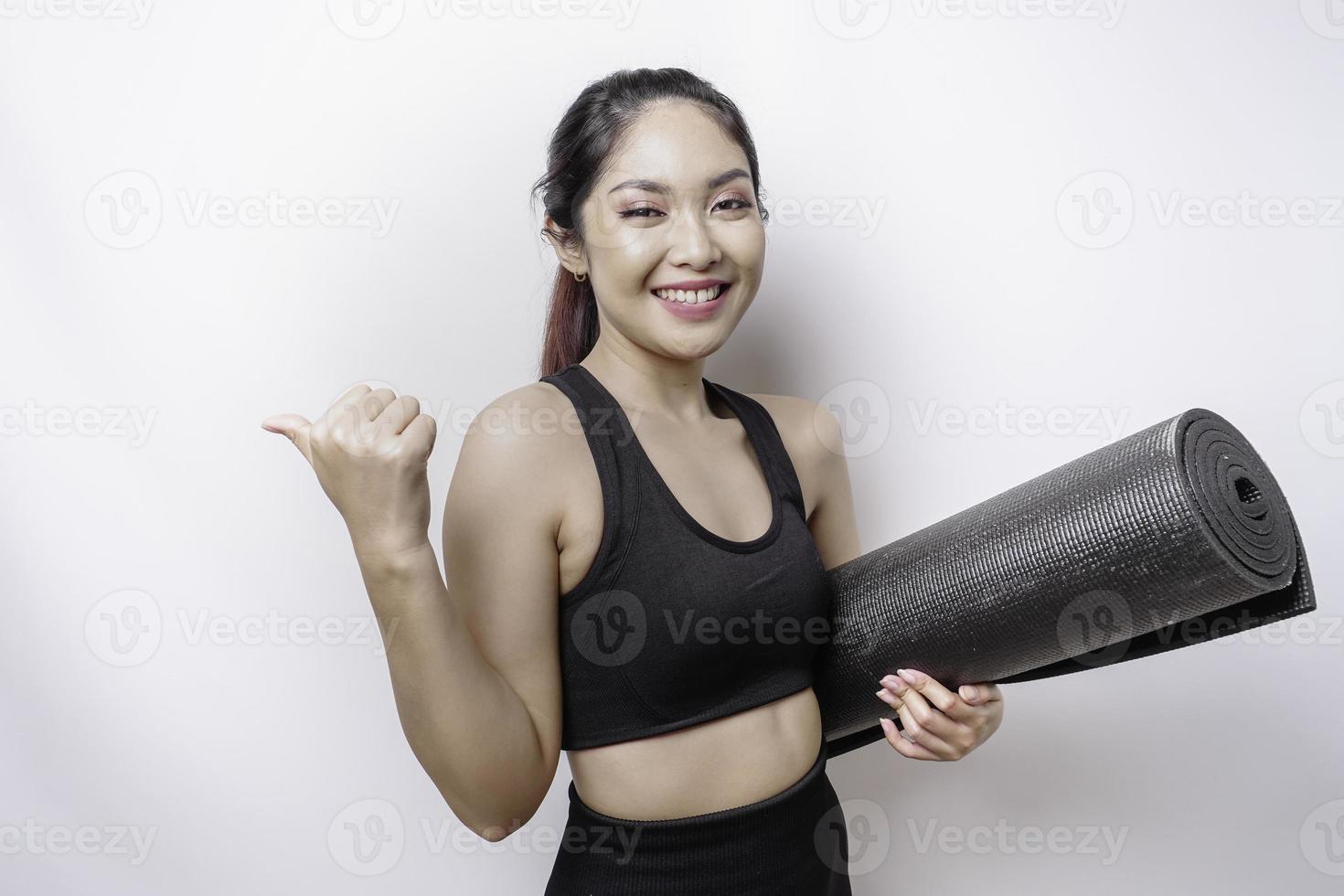 emocionada mujer asiática deportiva que usa ropa deportiva apuntando al espacio de la copia a su lado mientras lleva una alfombra de yoga, aislada de fondo blanco foto