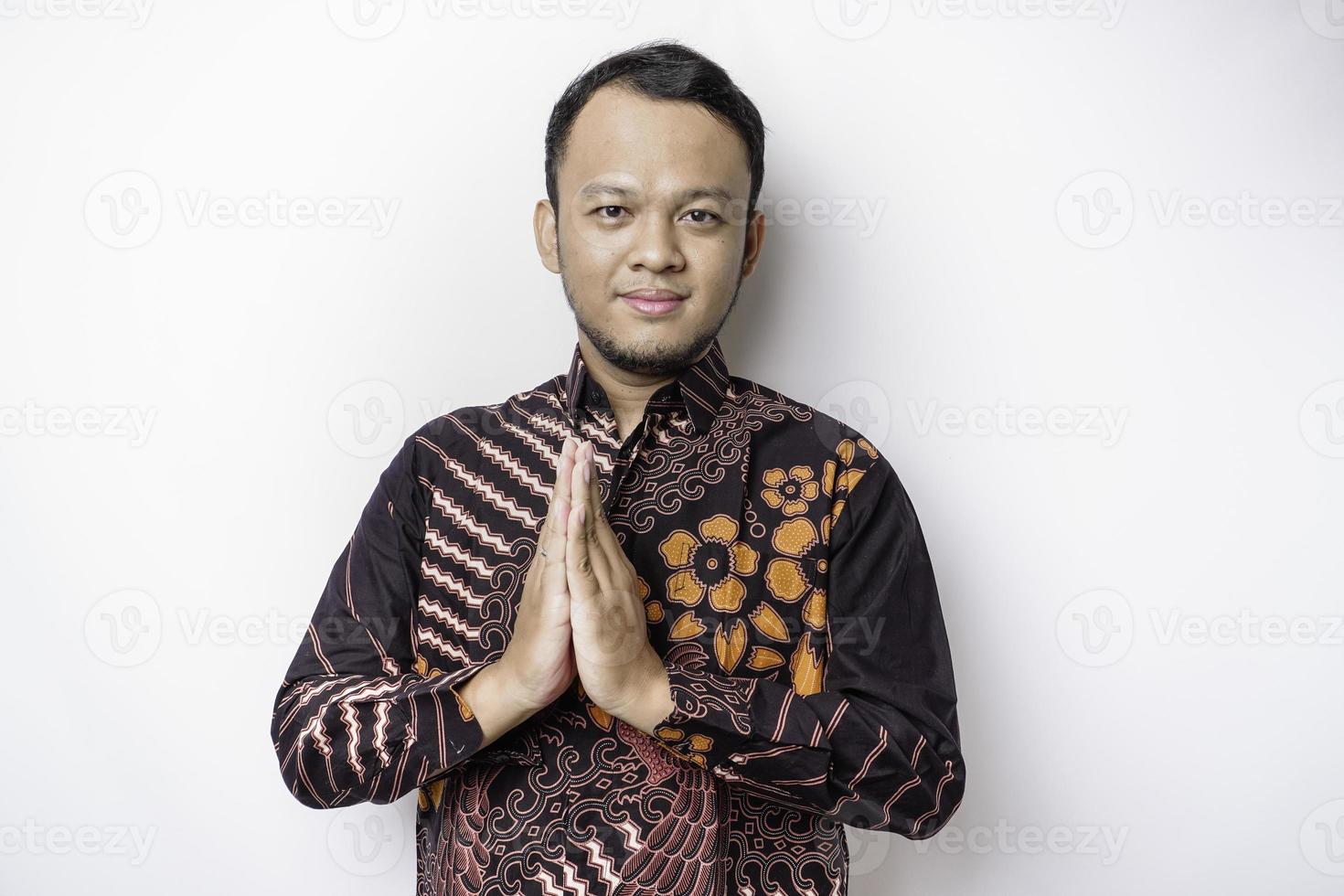 Smiling young Asian man wearing batik shirt, gesturing traditional greeting isolated over white background photo