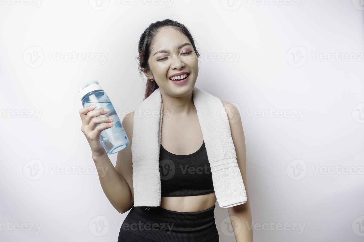 mujer asiática deportiva posando con una toalla en el hombro y sosteniendo una botella de agua, sonriendo y relajándose después del entrenamiento foto