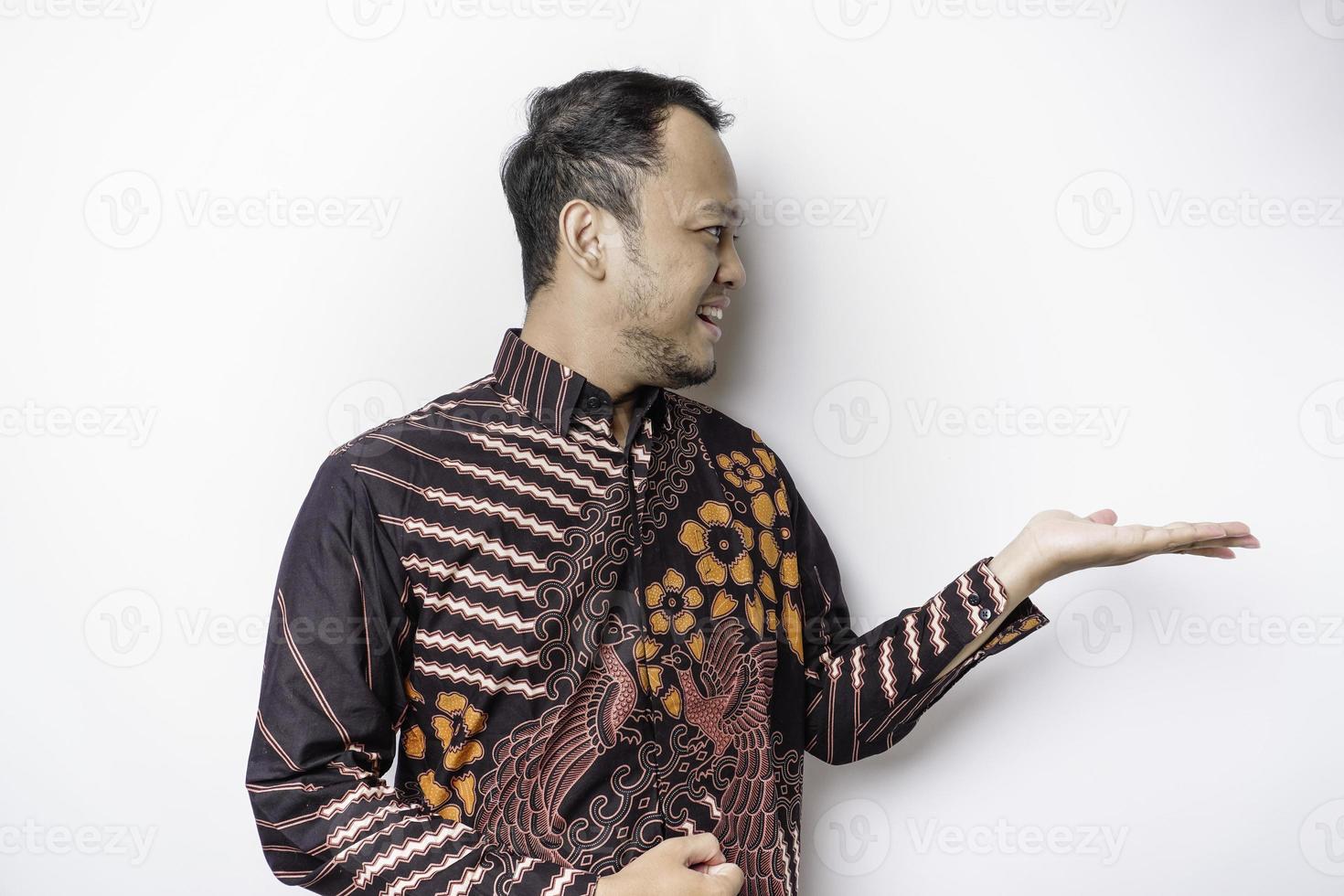 Excited Asian man wearing batik shirt pointing at the copy space beside him, isolated by white background photo