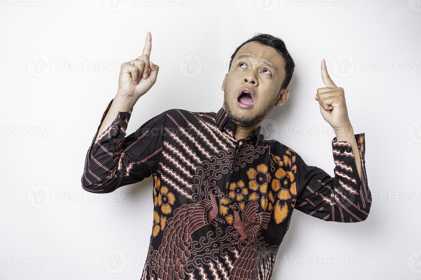 sorprendido hombre asiático con pantalones batik apuntando al espacio de copia encima de él, aislado por fondo blanco foto