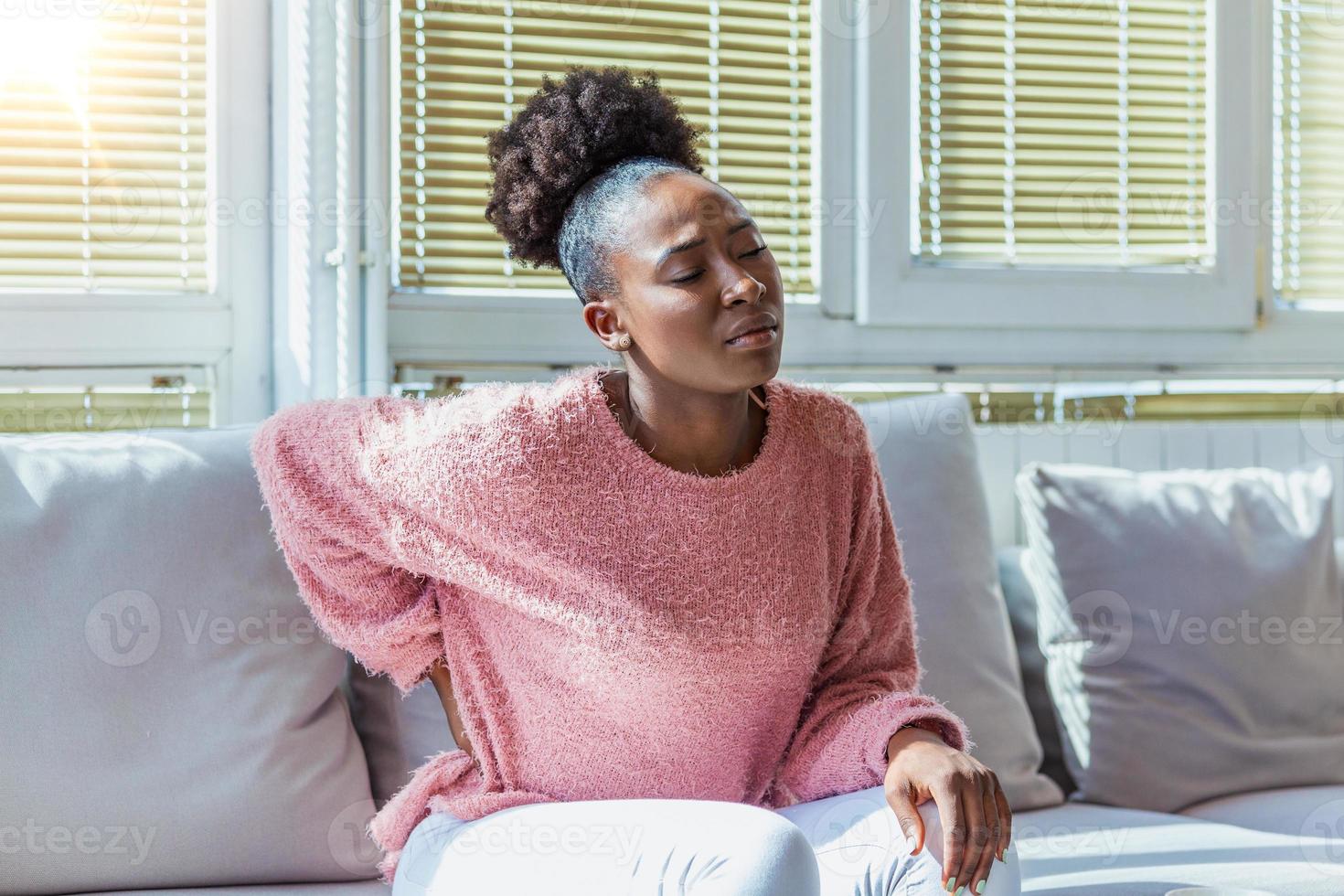 joven negra que sufre de dolor de espalda en casa. retrato de una joven morena sentada en el sofá de casa con dolor de cabeza y dolor de espalda. hermosa mujer con dolor de riñón o espinal foto