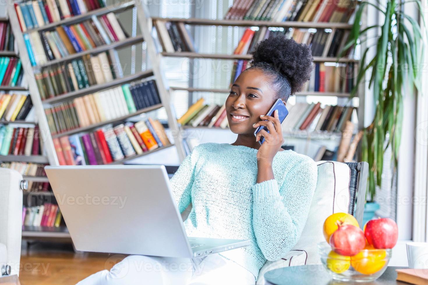 Happy woman sitting on sofa with laptop and talking on phone at home. Young successful businesswoman working from home while talking at phone. College student studying on laptop and using phone. photo