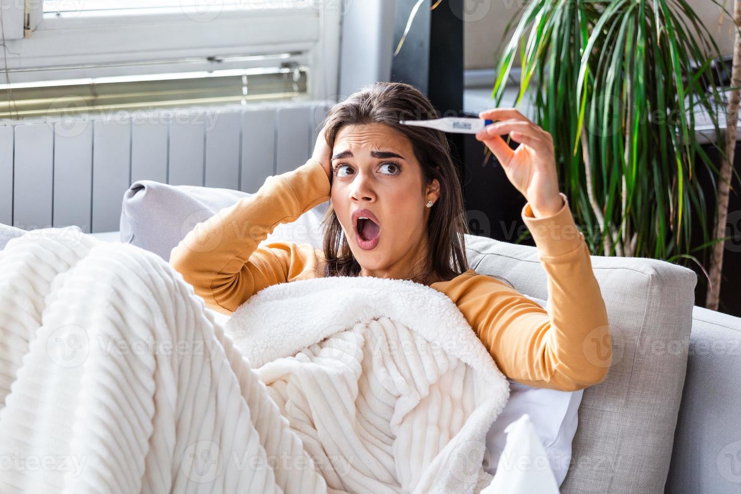 Sickness, seasonal virus problem concept. Woman being sick having flu lying on sofa looking at temperature on thermometer. Sick woman lying in bed with high fever. Cold flu and migraine. photo