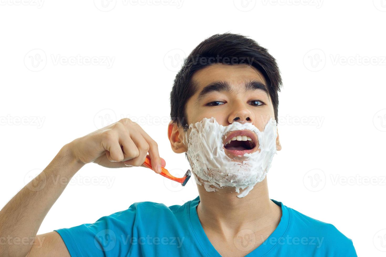 young cute guy with foam on his face opened his mouth and shaves his beard machine close-up photo