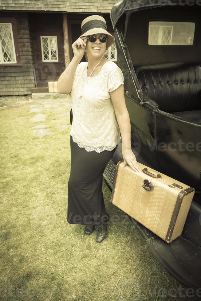 feliz chica vestida de los años 20 sosteniendo una maleta junto a un coche antiguo foto