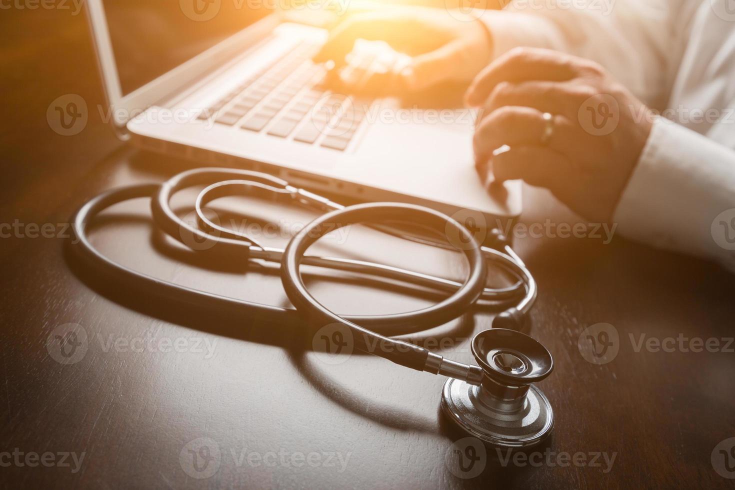 Medical Stethoscope Resting on Desk As Male Hands Type on Computer Keyboard. photo
