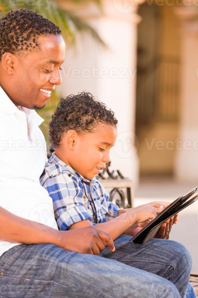 African American Father and Mixed Race Son Using Computer Tablet on Bench in Park photo