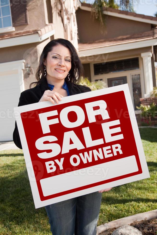 mujer hispana sosteniendo a la venta por el propietario letrero de bienes raíces frente a la casa foto