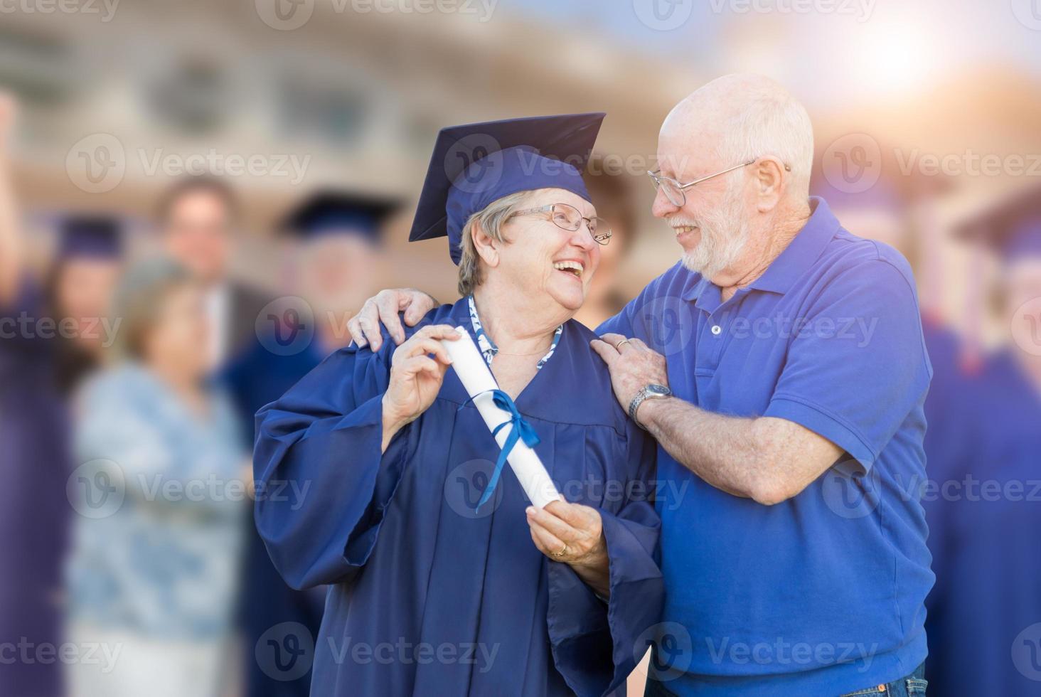 Senior Adult Woman In Cap and Gown Being Congratulated By Husband At Outdoor Graduation Ceremony. photo