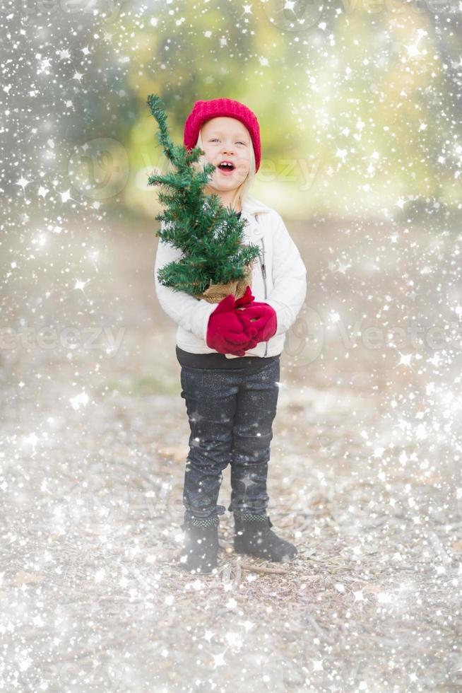 niña en mitones sosteniendo un pequeño árbol de navidad con efecto de nieve foto