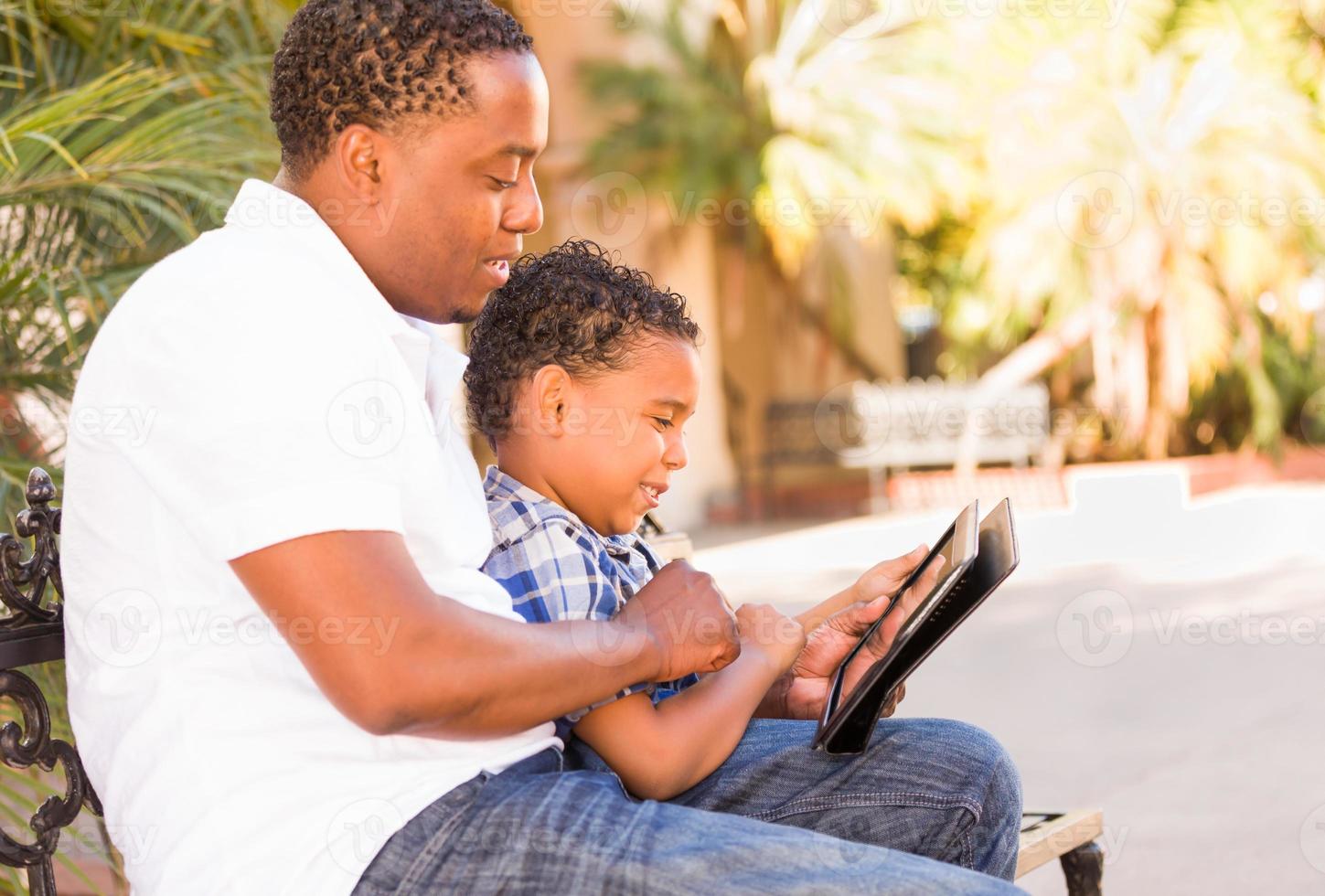 African American Father and Mixed Race Son Using Computer Tablet on Bench in Park photo