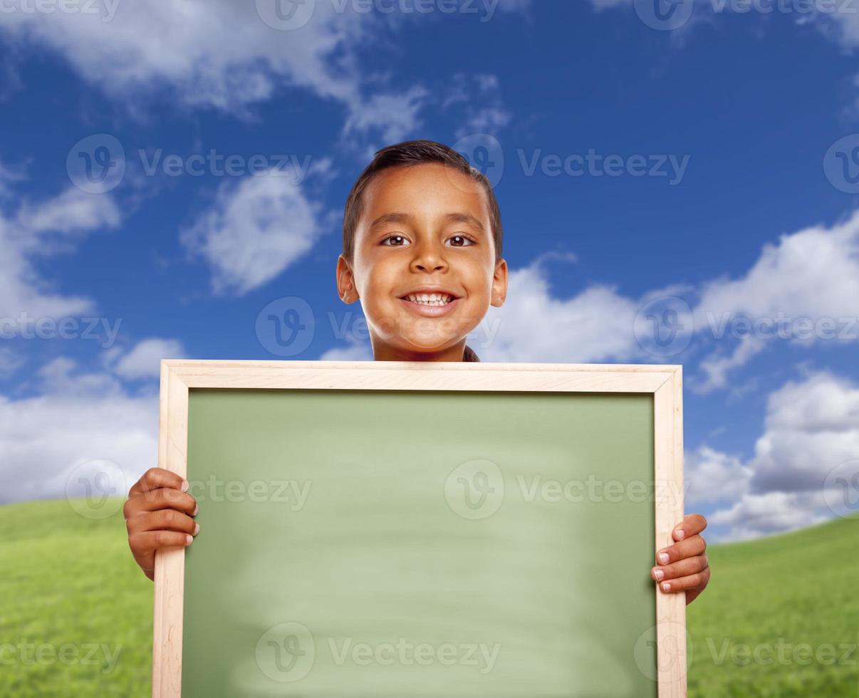 niño hispano feliz en el campo de hierba sosteniendo una pizarra en blanco foto