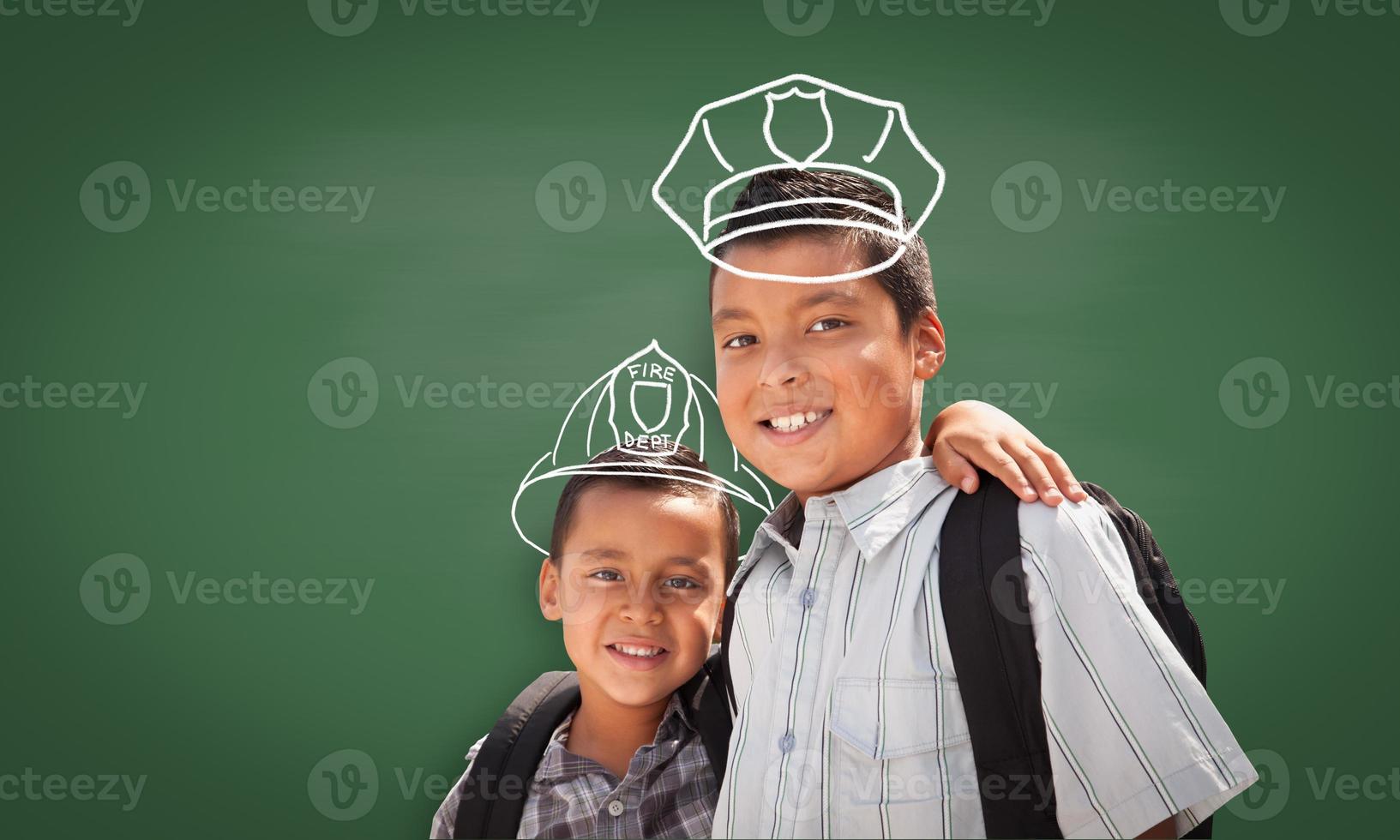 Young Hispanic Student Boy Wearing Backpack Front Of Blackboard with Fireman Helmet And Policeman Hat Drawn In Chalk Over Heads photo