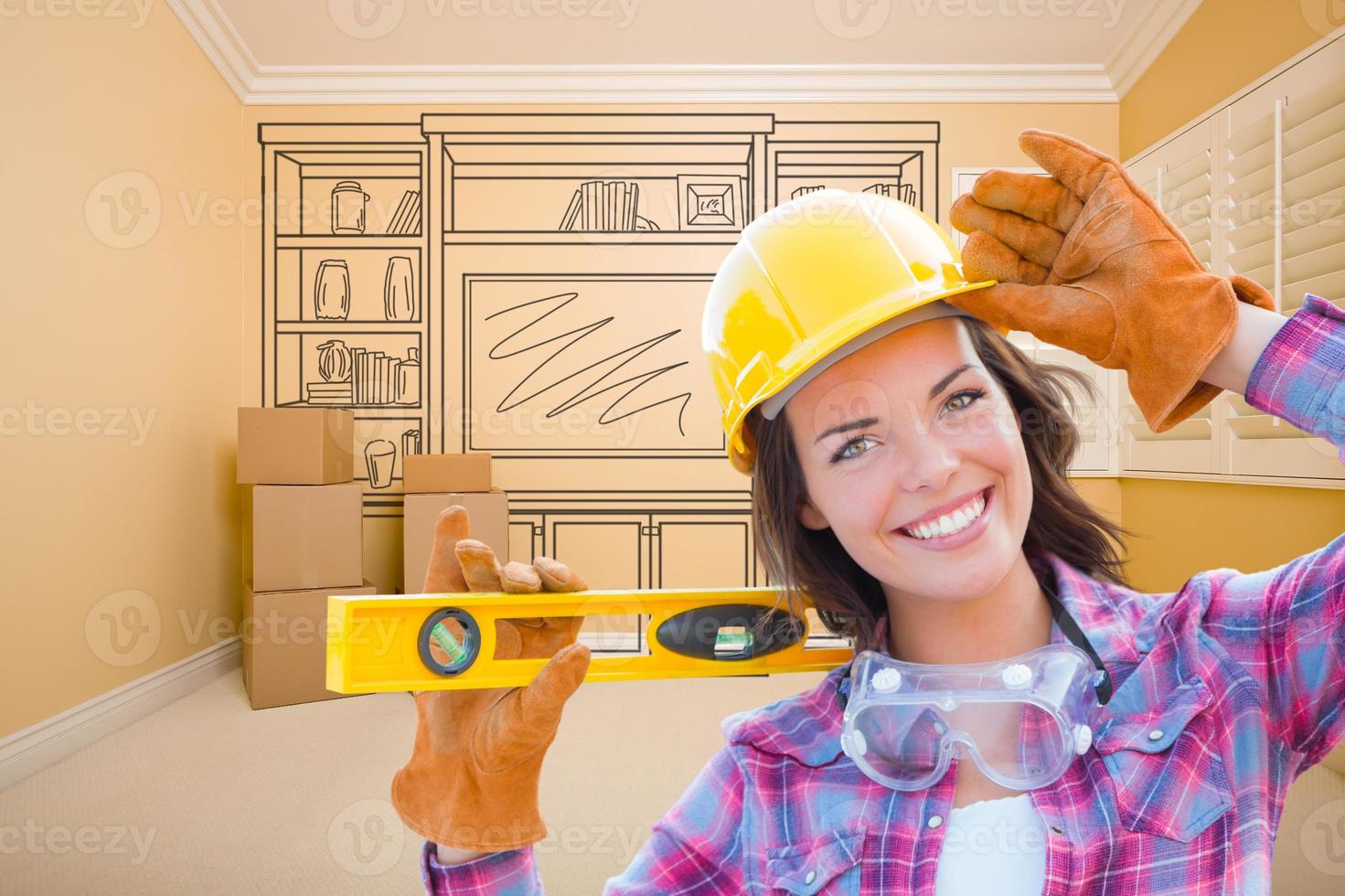 Female Construction Worker Holding Level In Front of Custom Built In Entertainment Unit Drawing in Empty Room. photo