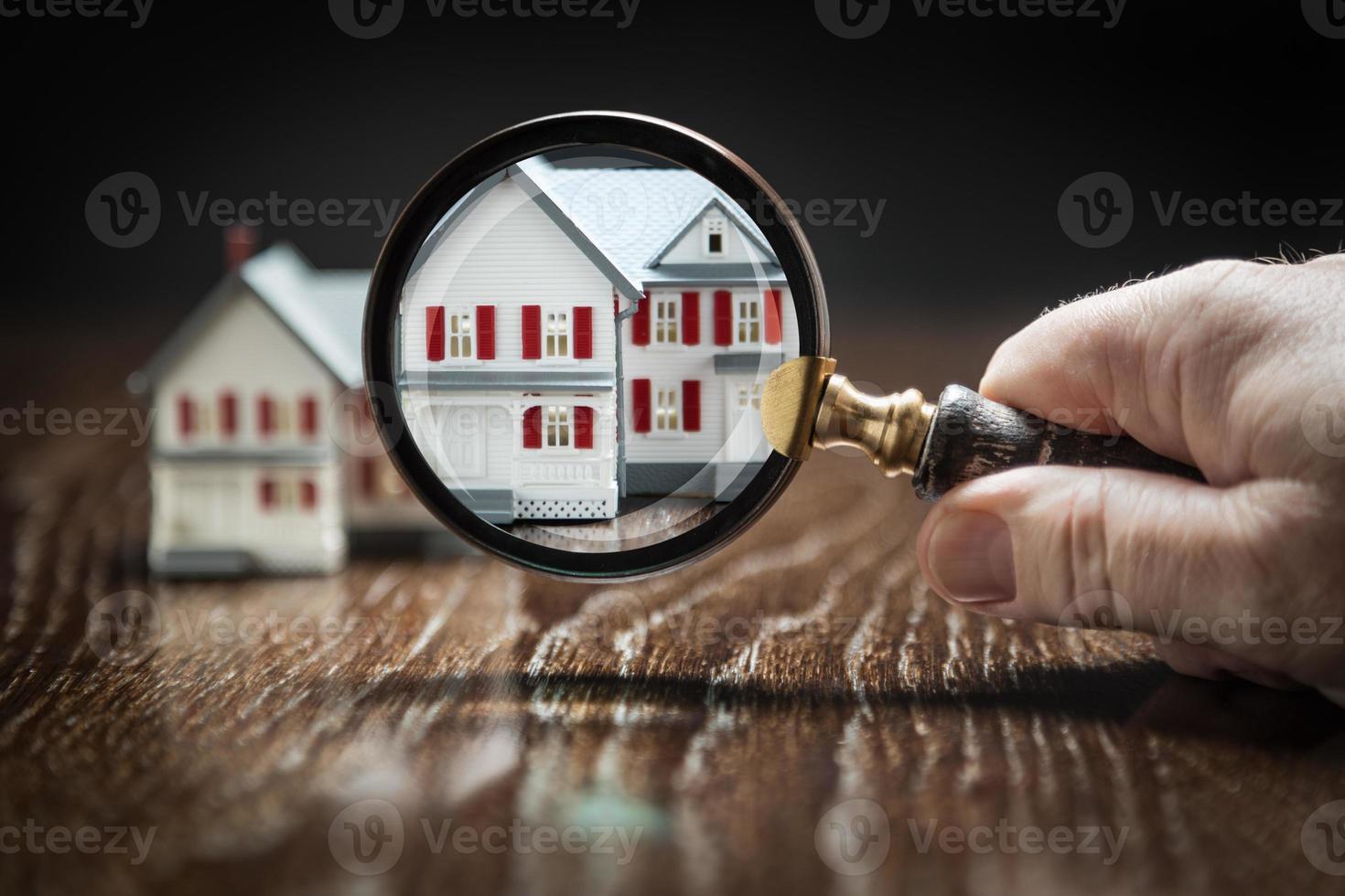 Hand Holding Magnifying Glass Up To Model Home on Reflective Wooden Surface. photo
