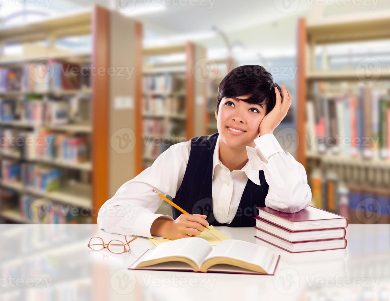 Young Female Mixed Race Student With Books and Paper Daydreaming In Library Looking To the Left. photo