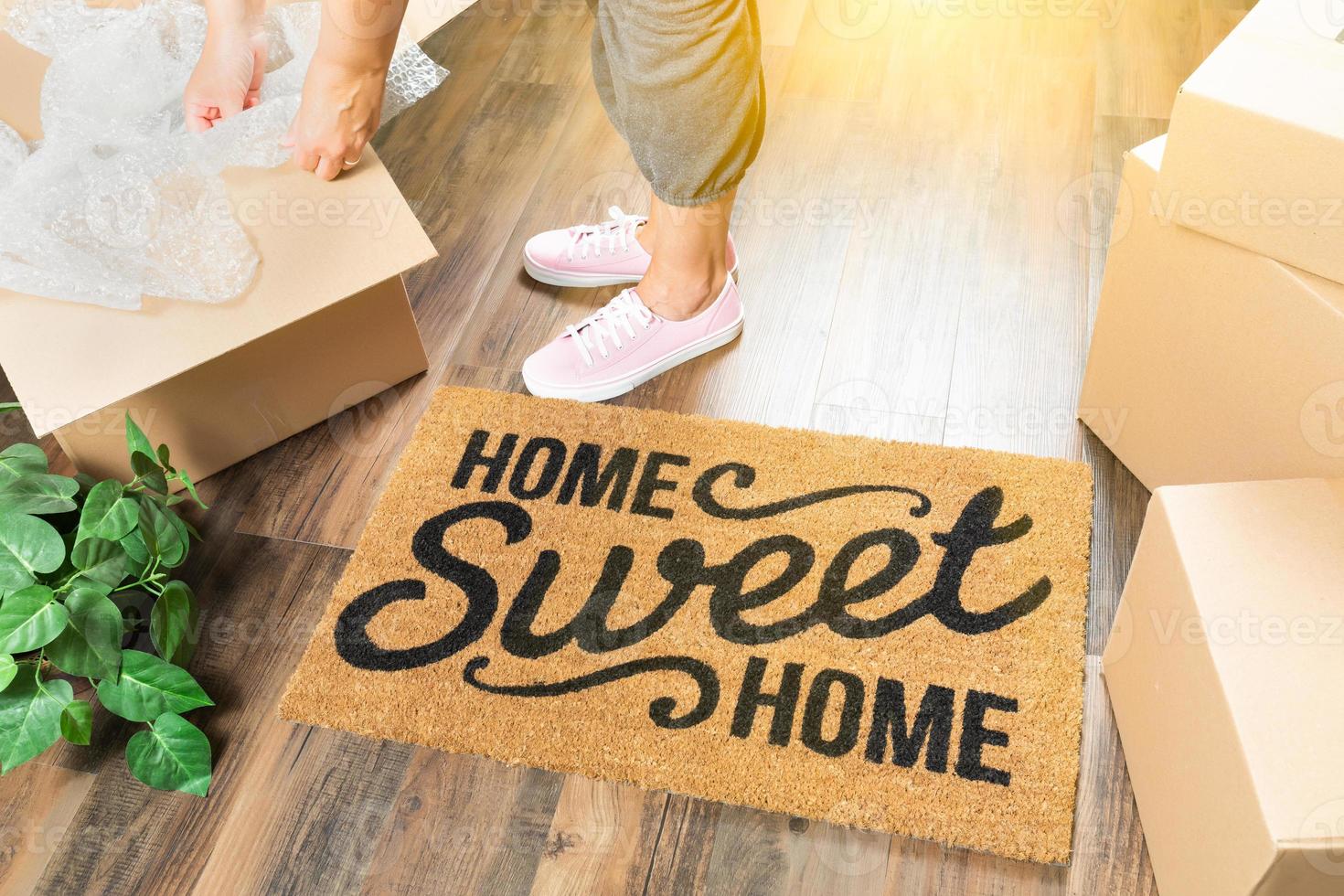 Woman in Pink Shoes and Sweats Unpacking Near Home Sweet Home Welcome Mat, Moving Boxes and Plant photo