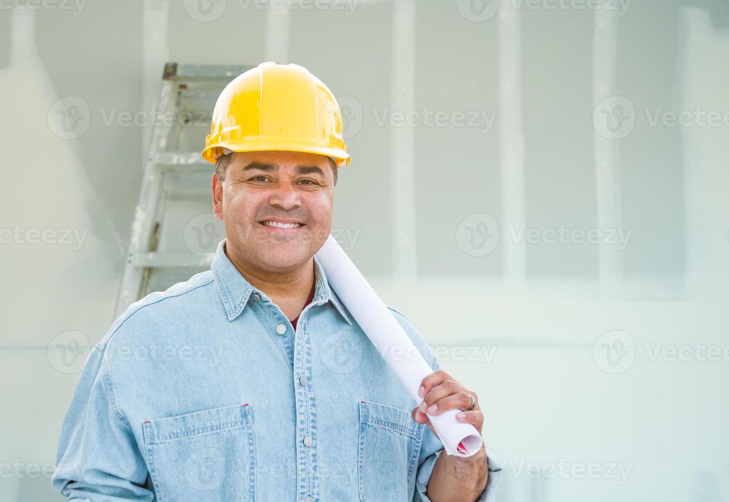 Hispanic Male Contractor with Blueprint Plans Wearing Hard Hat In Front of Drywall and Ladder photo