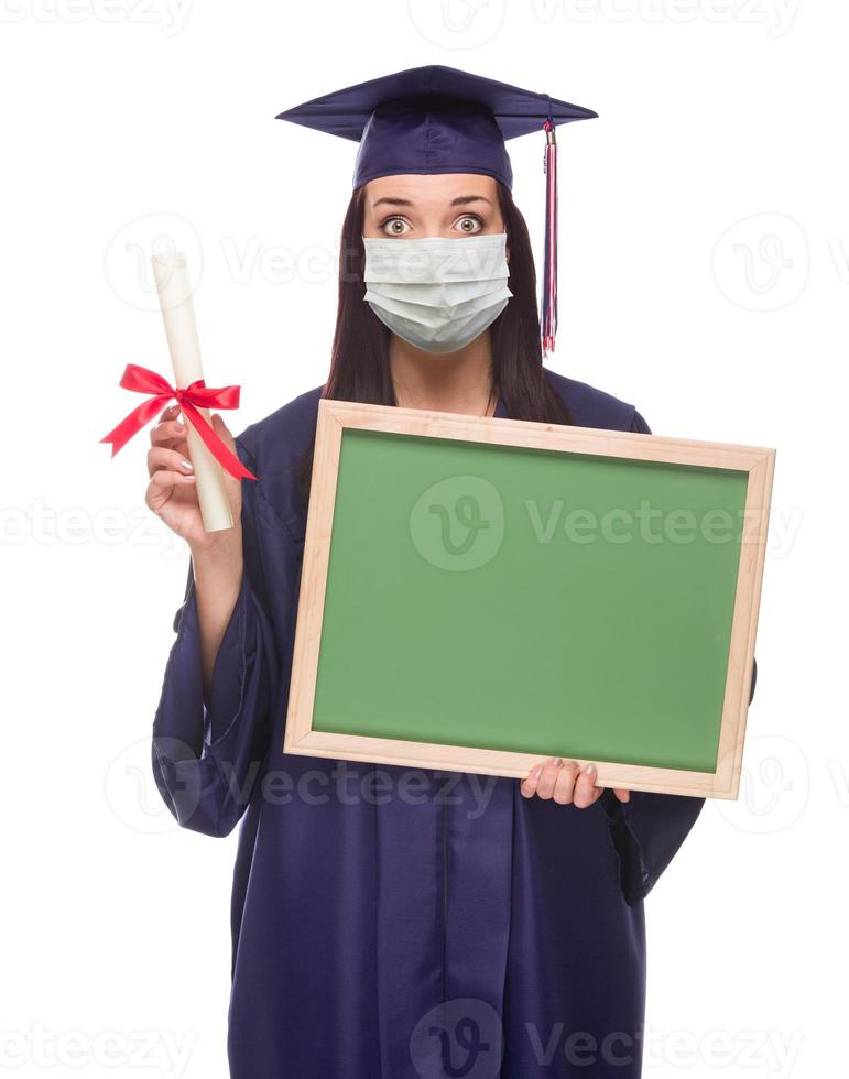 mujer graduada con mascarilla médica y toga y birrete sosteniendo una pizarra en blanco aislada en un fondo blanco foto
