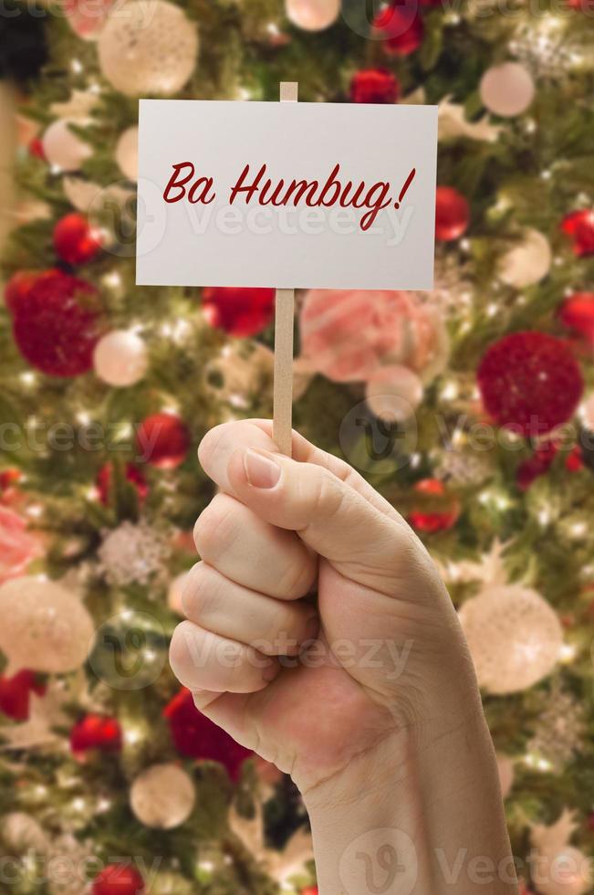 mano que sostiene la tarjeta ba humbug frente al árbol de navidad decorado. foto