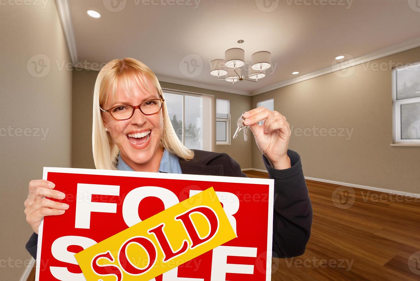 mujer joven atractiva con llaves nuevas y cartel de bienes raíces vendidos en una habitación vacía de la casa foto