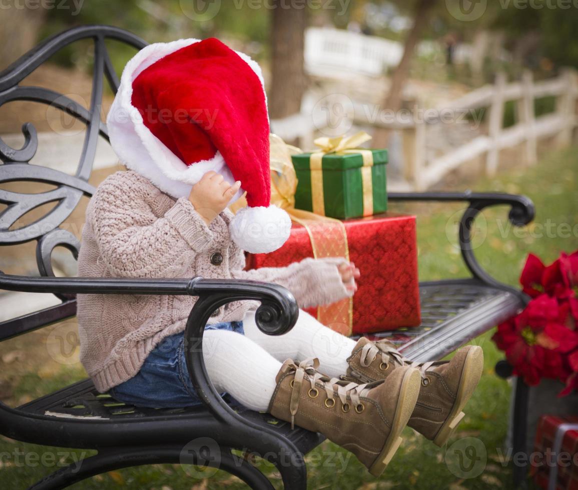 Young Child Wearing Santa Hat Sitting with Christmas Gifts Outside. photo