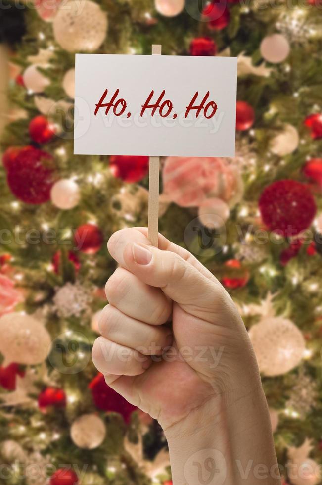 mano sosteniendo la tarjeta ho ho ho frente al árbol de navidad decorado. foto