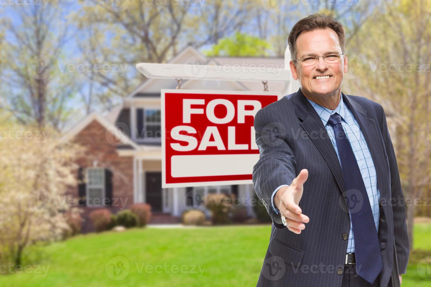 agente masculino alcanzando un apretón de manos frente a la casa y en venta signo de bienes raíces foto