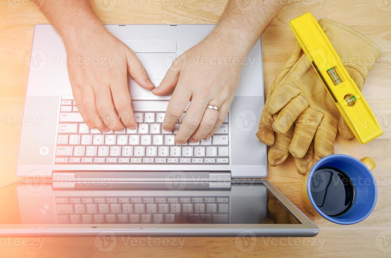 Work Gloves, Level and Coffee Cup Next To Contractor Reviewing Project on Laptop photo